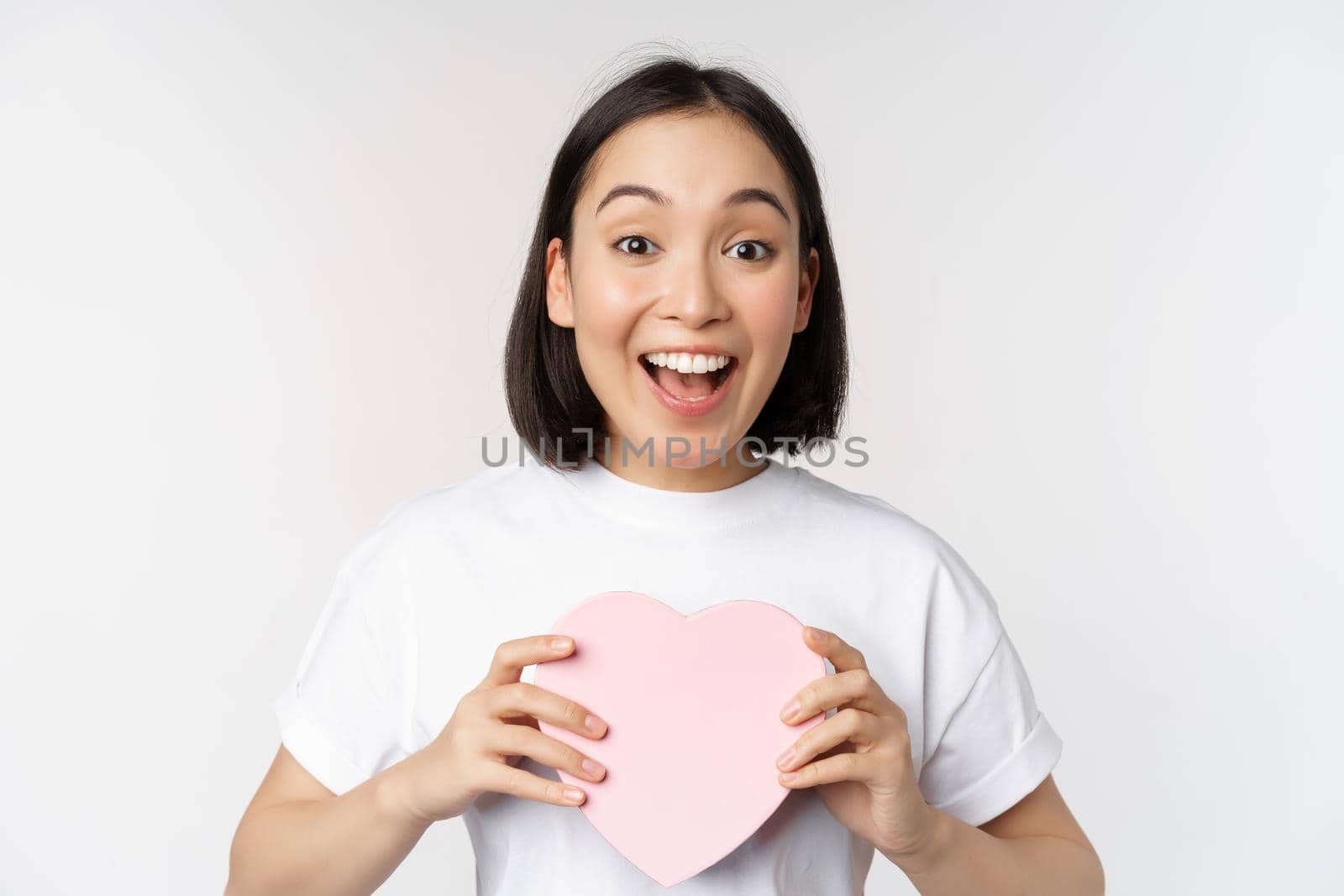 Valentines day. Happy asian girl receive romantic gift, holding heart shaped box and smiling excited, standing over white background.