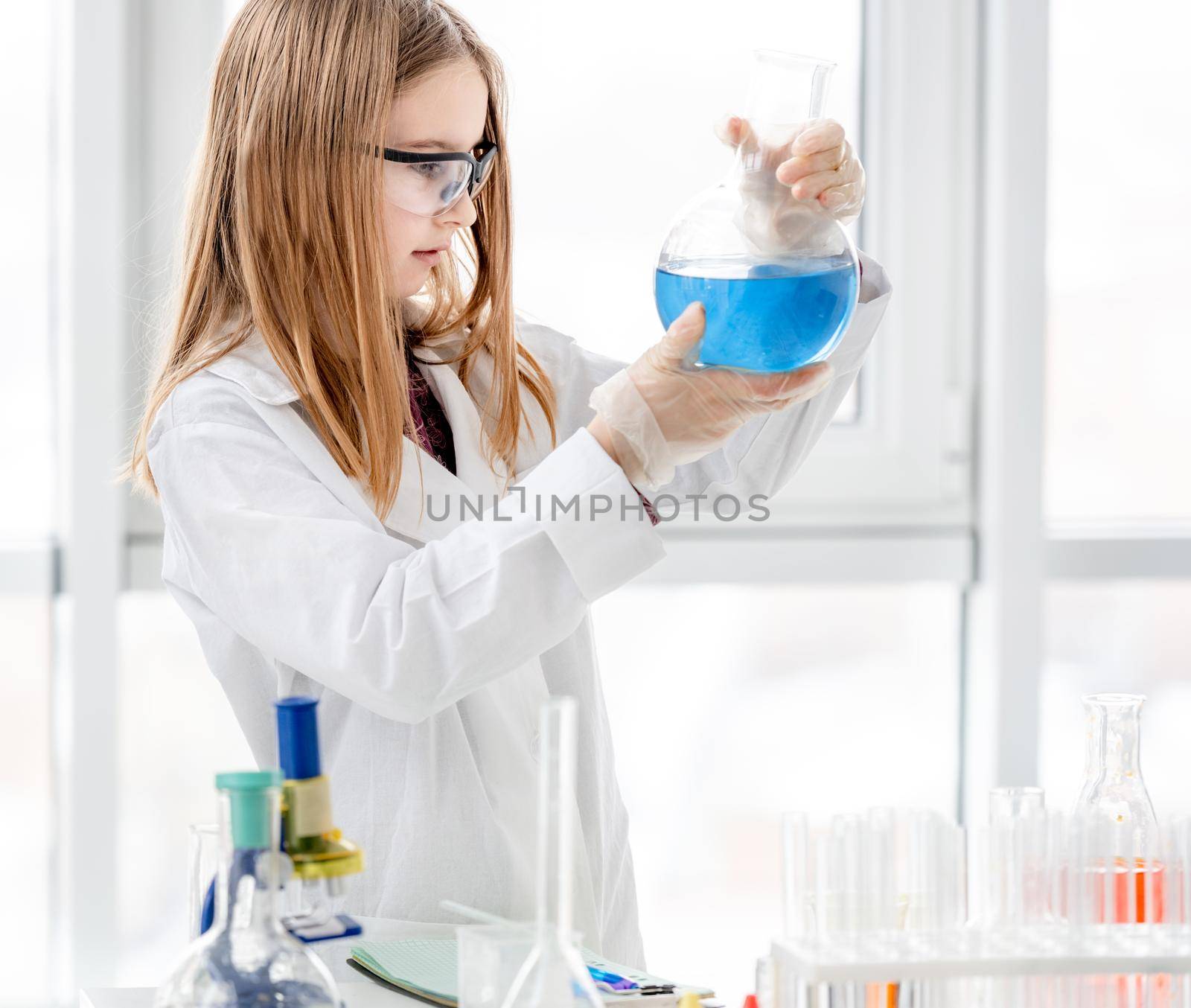 Smart girl doing scientific chemistry experiment wearing protection glasses, holding bottle and measuring blue liquid. Schoolgirl with chemical equipment on school lesson