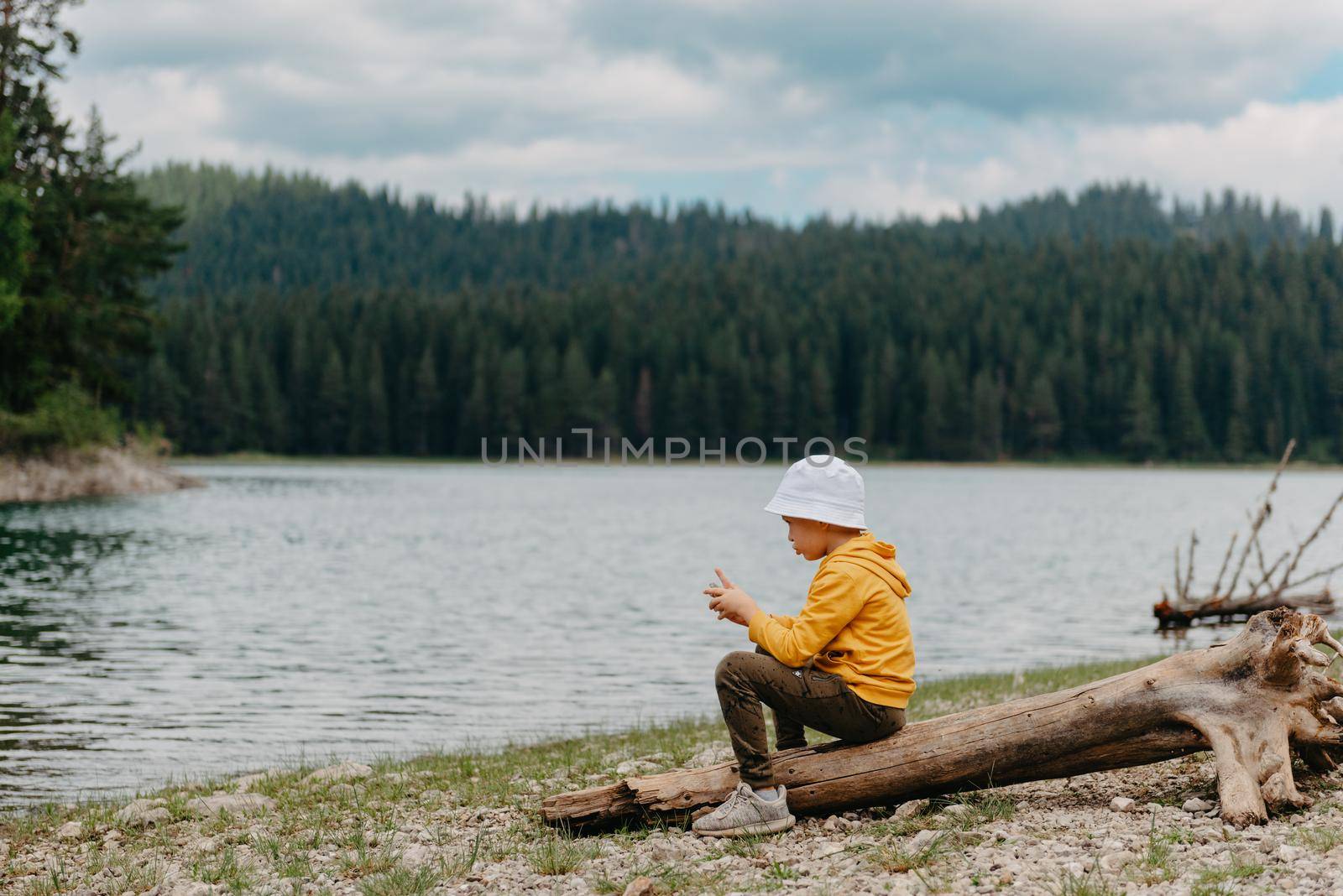 Little boy sitting on the log near the mountain Lake. A lonely boy sits quietly and looks ahead at a beautiful view of mountains. little boy lake mn, little boy lake resort.