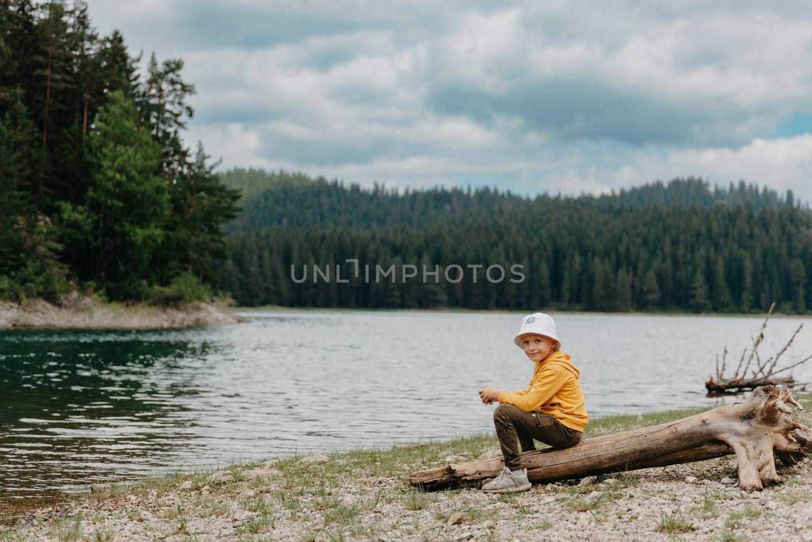 Little boy sitting on the log near the mountain Lake. A lonely boy sits quietly and looks ahead at a beautiful view of mountains. little boy lake mn, little boy lake resort by Andrii_Ko