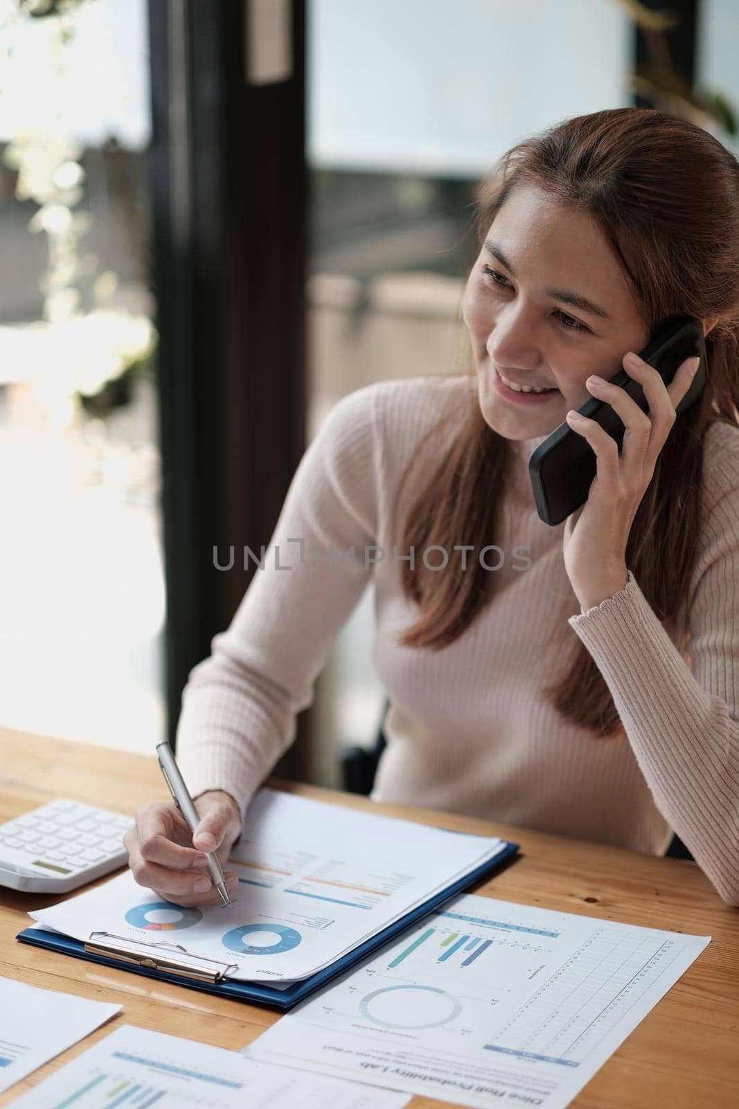 Portrait of young asian woman sitting at her desk working with calculator and paperwork while calling mobile phone in modern office
