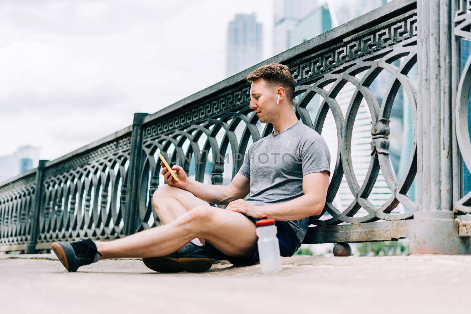 Tired Young man runner sitting on road, using mobile phone and relaxing after sport training. Holding water bottle while doing fitness workout in city urban street, cloudy sky at summer.
