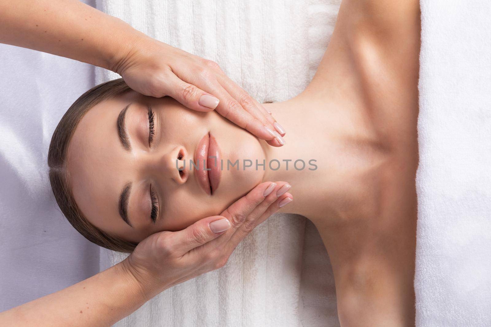 Releasing stress. Top view of beautiful young woman lying on back while massage therapist massaging her face