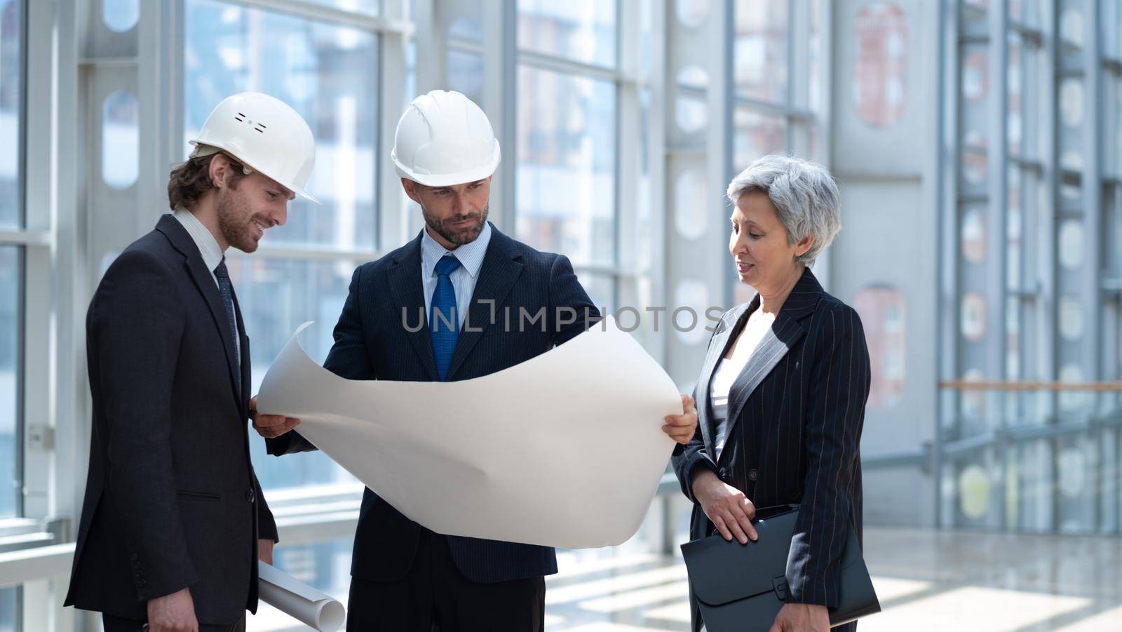 Multi ethnic business architect people in safety helmets discussing blueprint standing in the lobby at office