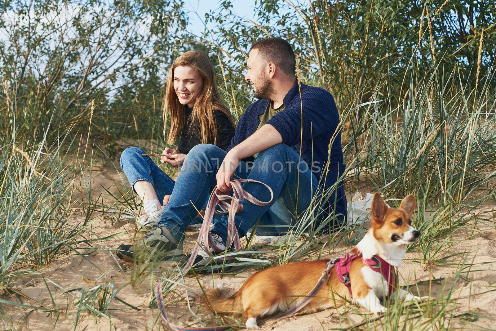 Young happy couple of man and woman with corgi dog siting at sand. Two persons with pet enjoying beautiful sunny afternoon on beach