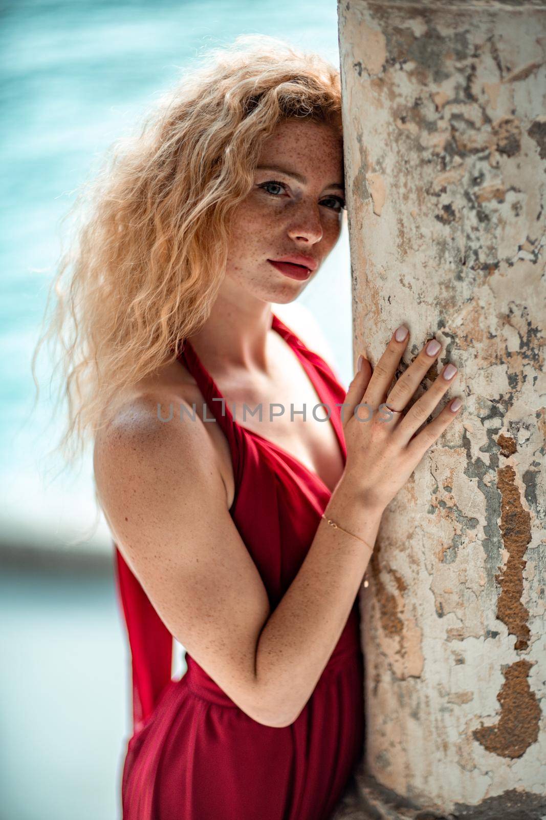 Outdoor portrait of a young beautiful natural redhead girl with freckles, long curly hair, in a red dress, posing against the background of the sea. by Matiunina