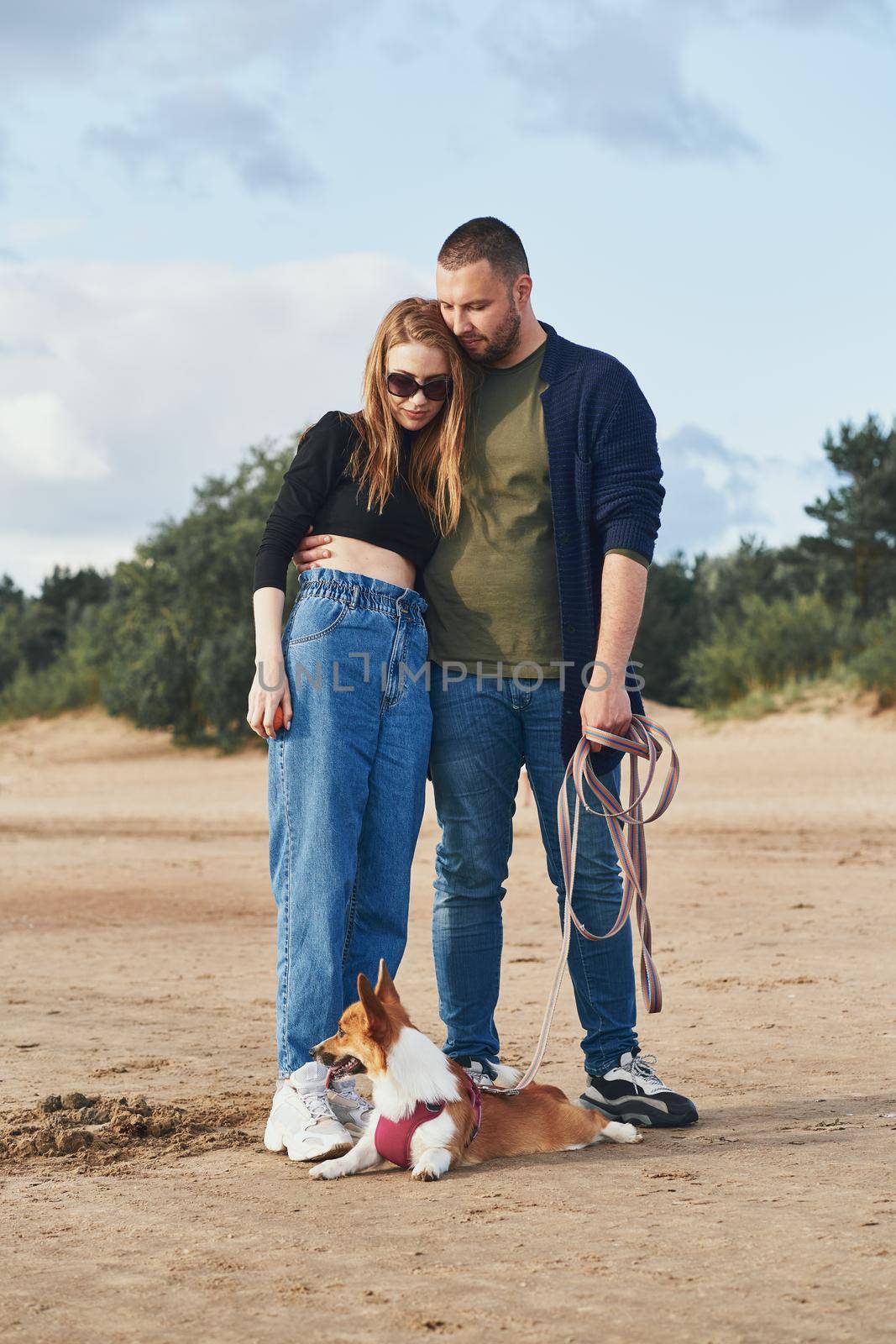 Young happy couple and dog standing on beach against pines and sand. Handsome man gently hugging woman while holding Corgi puppy on leash. Walking in summer nature of people in love