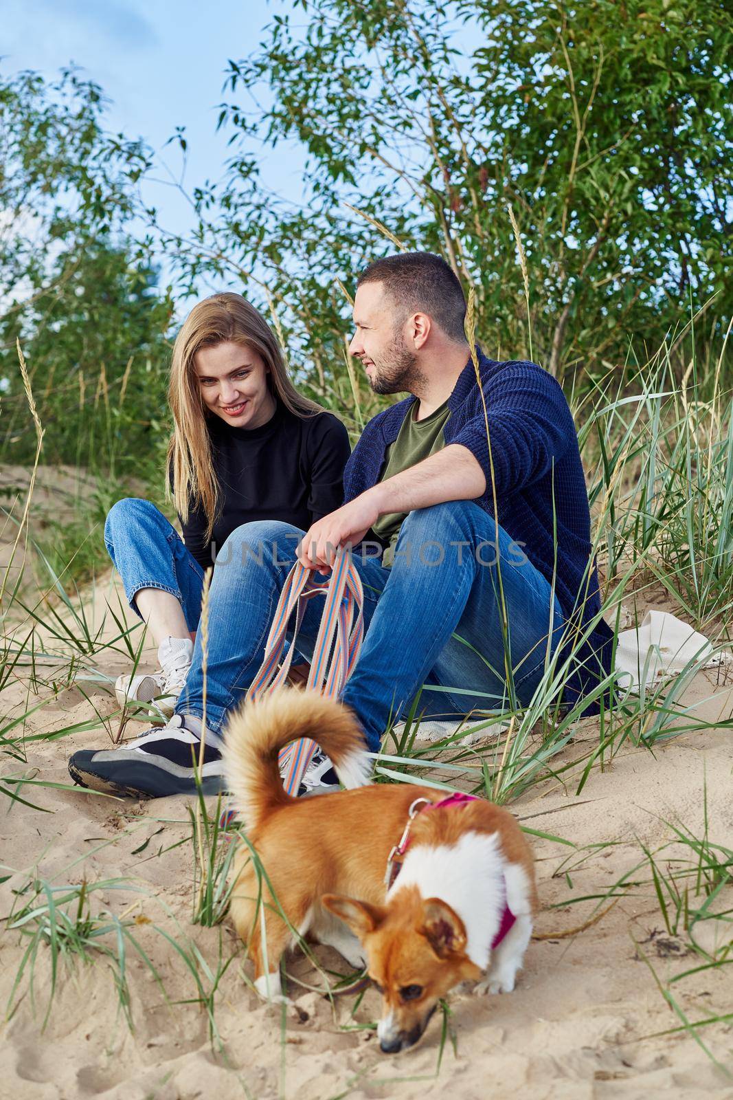 Young happy couple of man and woman with corgi dog sit at sand. Two persons in casual clothes by NataBene