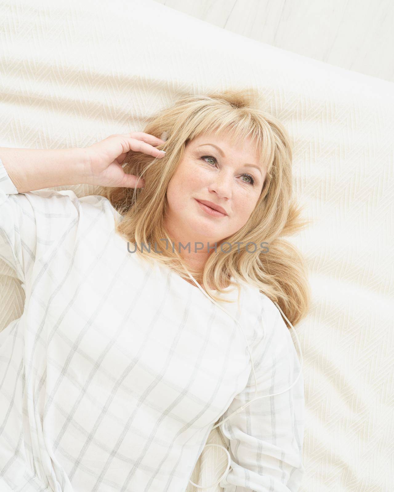 Mature woman with headphones relaxing at home, lying on bed and listening to music. Middle age beautiful blond female smiling and dreaming, self immersion