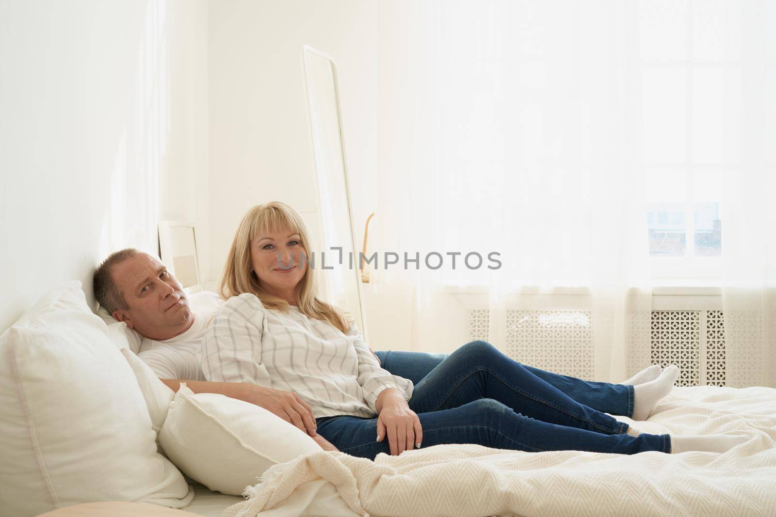 Full length portrait of mature couple in home interior on sofa. Handsome man and attractive middle age woman enjoying spending time together while lying in bed. People looking at camera