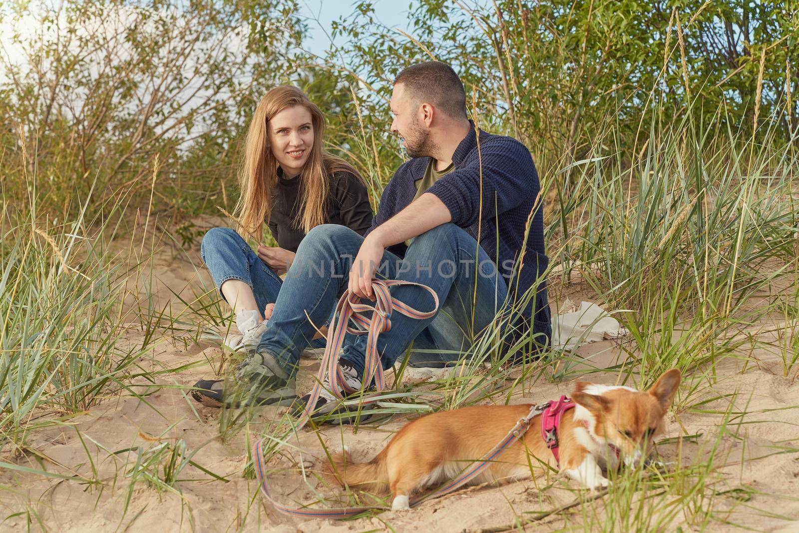 Young happy couple with corgi dog, man and woman siting at sand. Two persons in warm casual clothes talking in beautiful sunny afternoon on beach