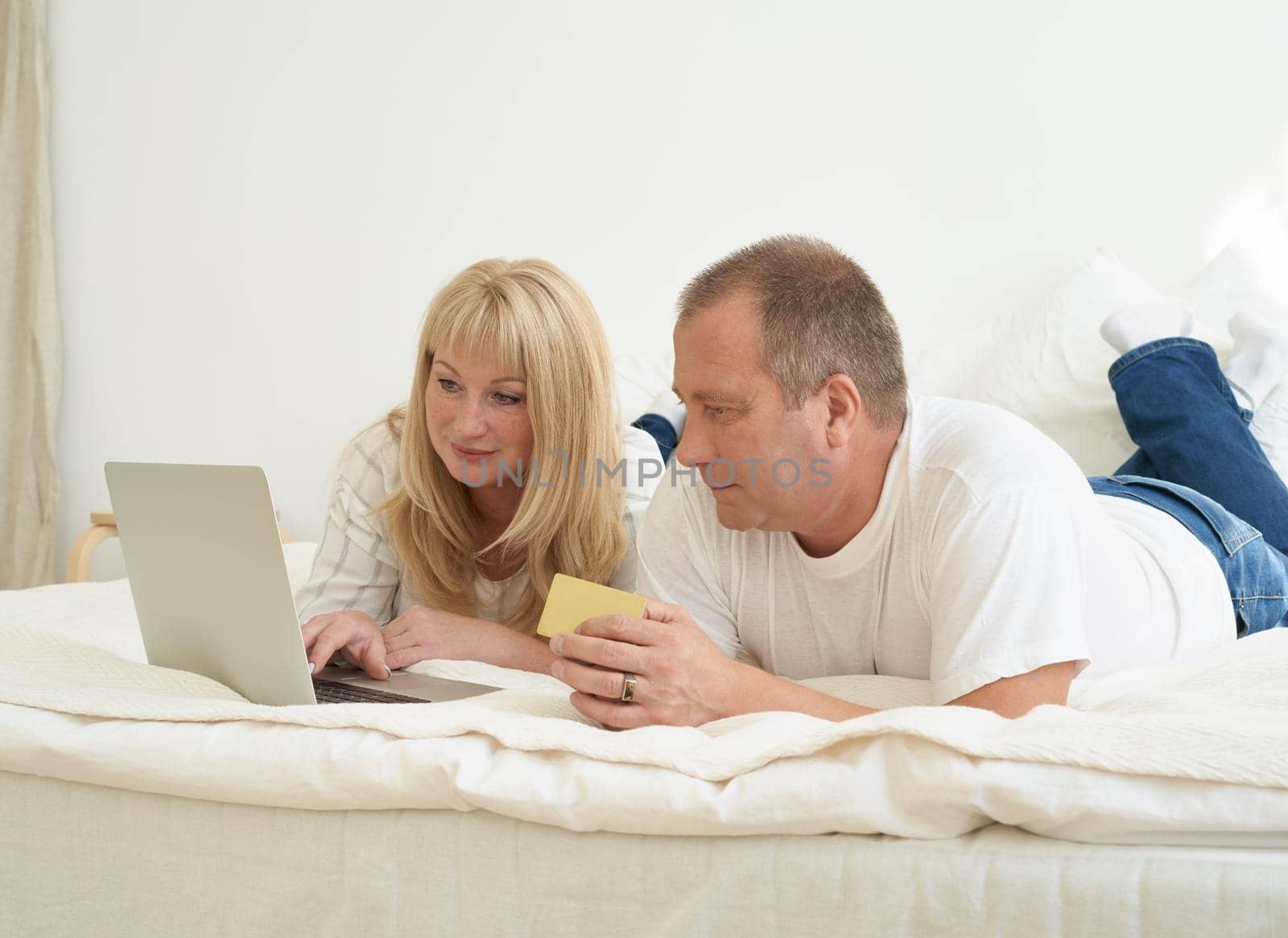 Mature family couple sitting at bed in modern home, looking at laptop screen. by NataBene