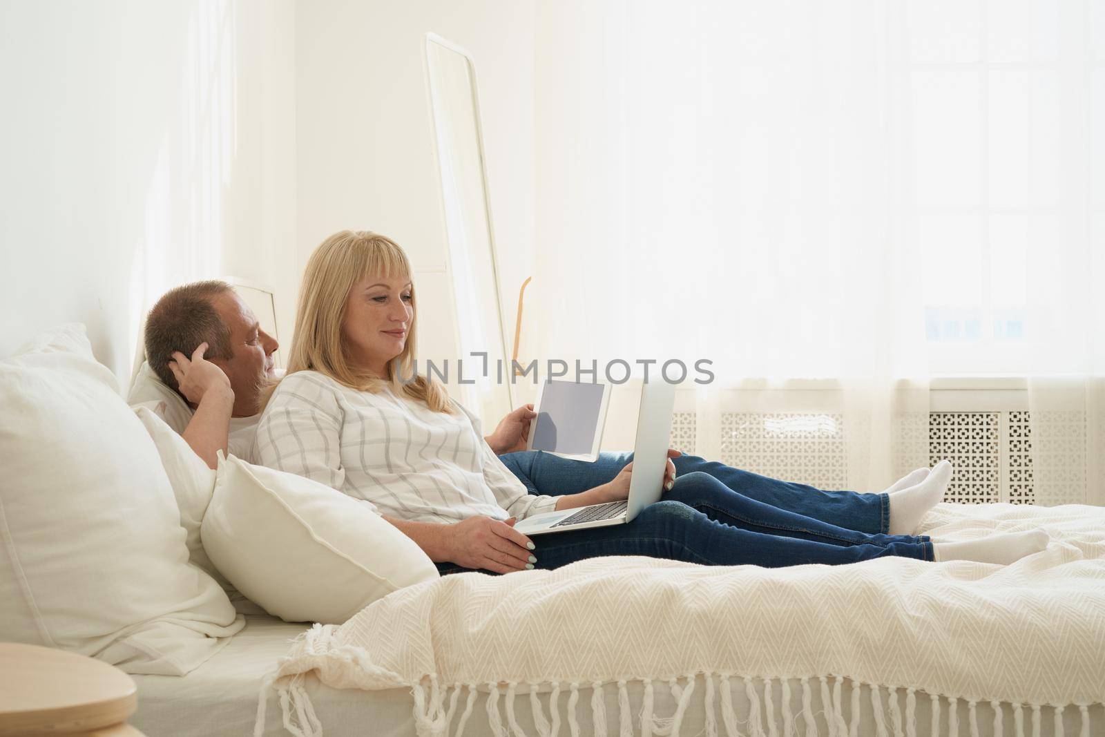 Full length portrait of mature couple with digital addiction at home. Handsome man with tablet and attractive middle age woman with laptop spending time together while lying in bed.