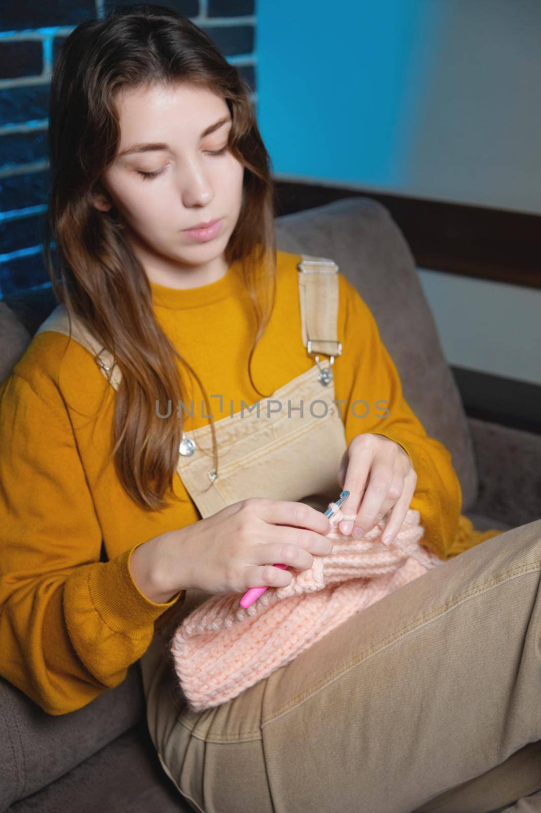 cute young caucasian woman sitting on sofa at home and crocheting wool product.