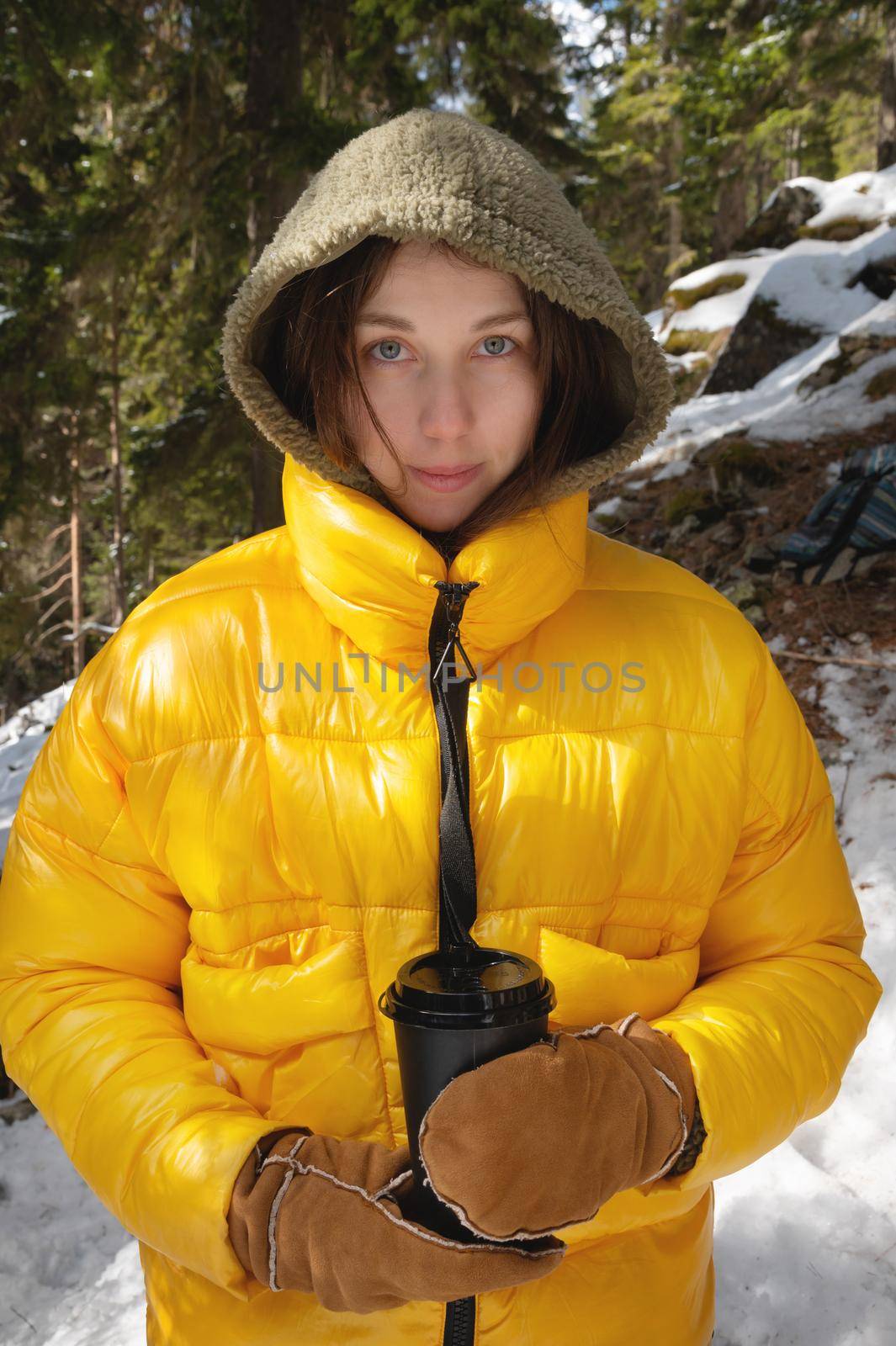 cute caucasian woman in a yellow jacket, in a hood and mittens, holds a paper glass with a hot drink in a coniferous mountain forest