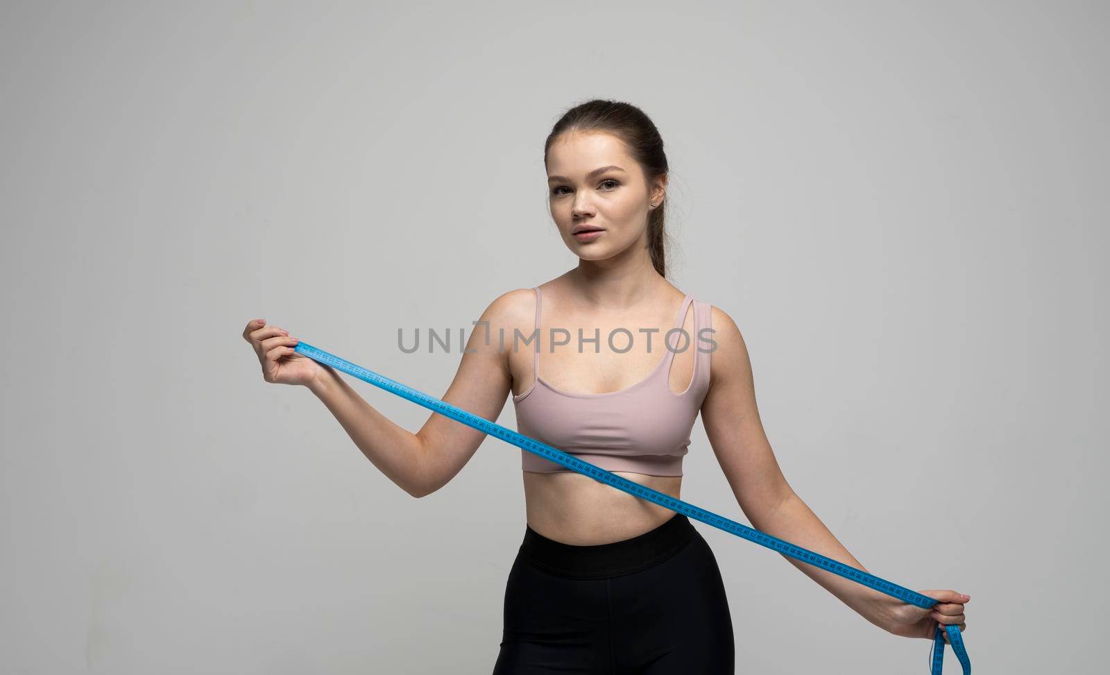 Weight loss and diet concepts. Attractive sporty slim brunette female in sportswear holding a blue measuring tape and looking in a camera. Health care and healthy nutrition