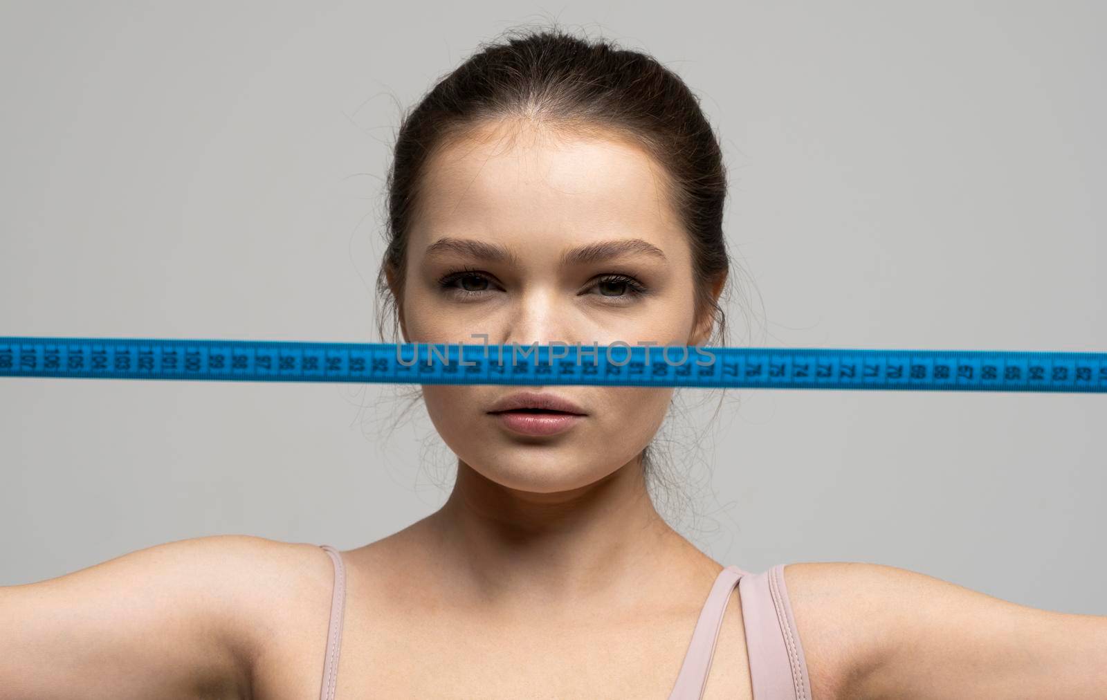 Close up portrait of attractive brunette woman with green eyes holding a blue measuring tape in front of her face and looking in a camera. Weight loss and diet concepts