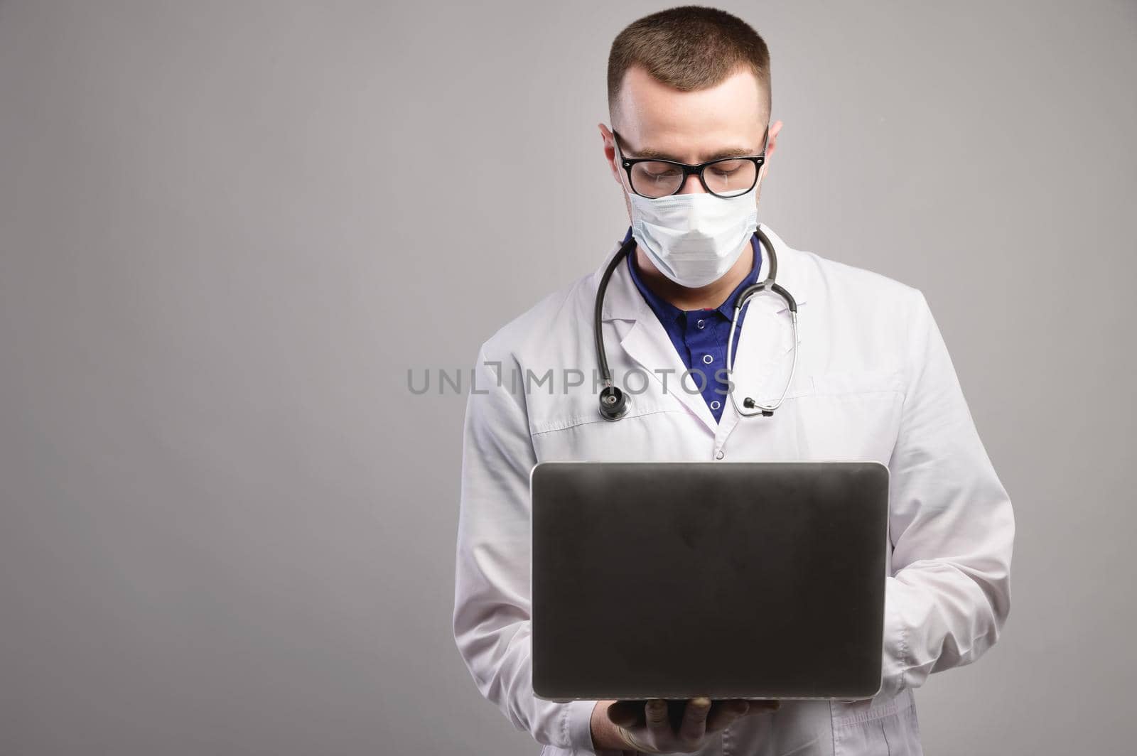 a young caucasian doctor in a mask and glasses uses his laptop. studio portrait on a gray background. a doctor in a white coat with a stethoscope around his neck looks into a laptop
