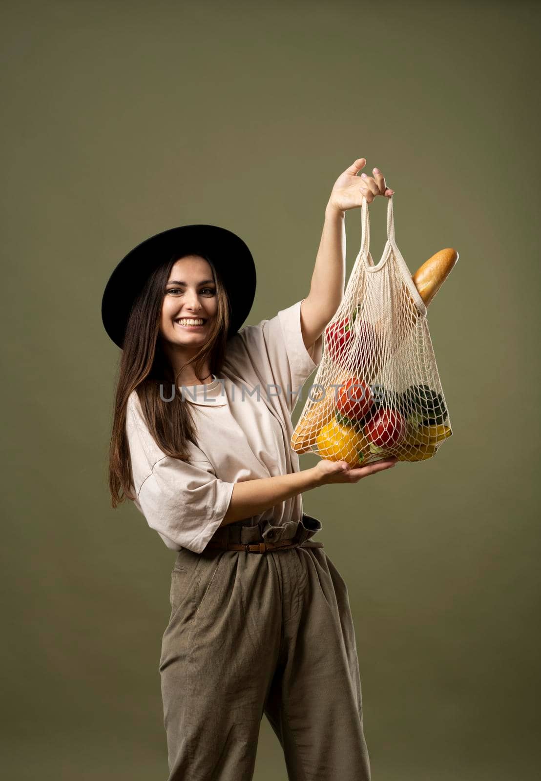 Cheerful brunette woman in beige t-shirt and black hat holding a mesh eco bag full of fruits and vegetables on natural green background in studio. Zero waste concept