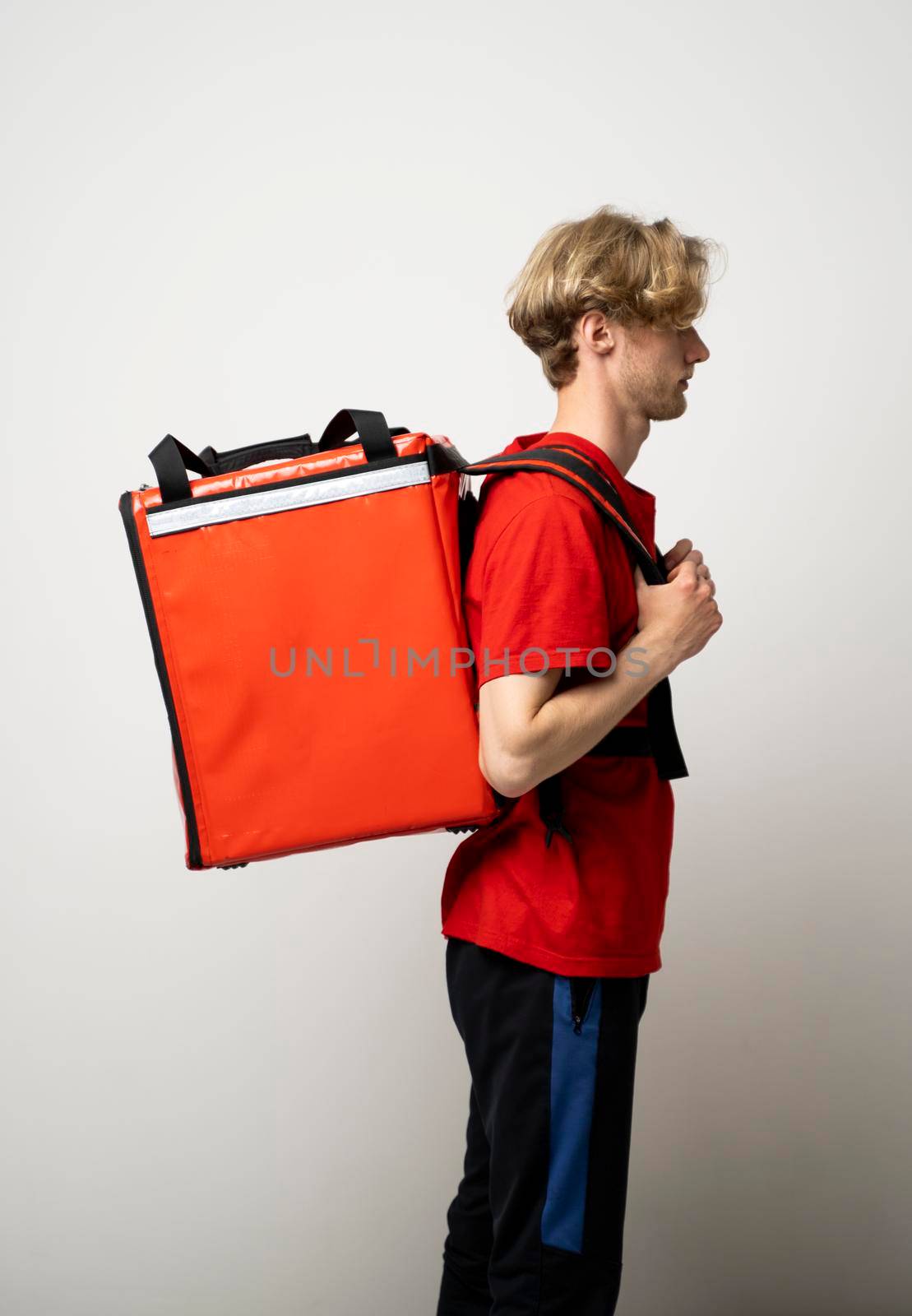 Fast Food Delivery Service Concept. Side view of smiling male courier wearing red uniform and holding thermal backpack bag isolated on studio background