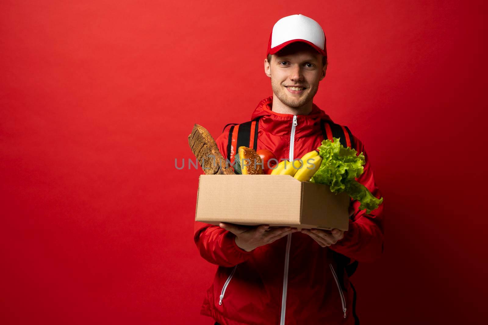 Happy satisfied delivery man, courier in a red uniform with a thermal bag on a shoulder delivered food to a customer at home. Online grocery shopping service concept