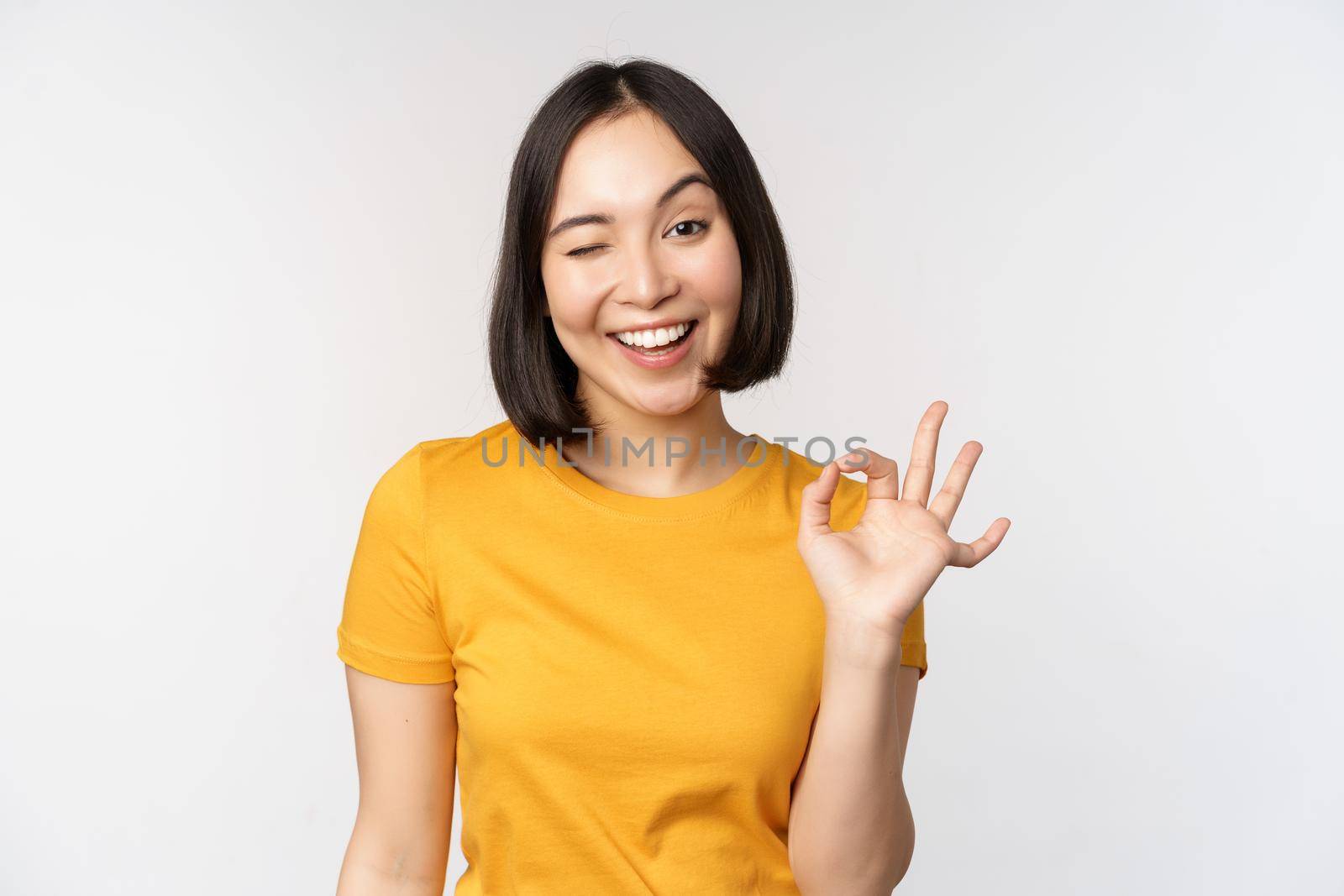 Beautiful young woman showing okay sign, smiling pleased, recommending smth, approve, like product, standing in yellow tshirt over white background.