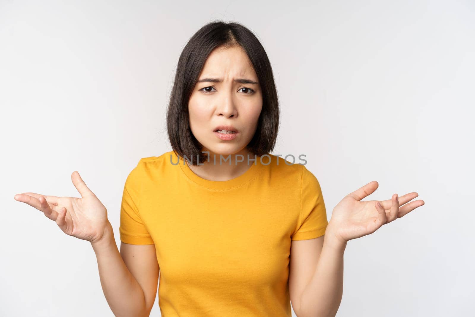 Frustrated, confused asian woman cant understand smth, looking clueless, shrugging shoulders, standing in yellow tshirt over white background.
