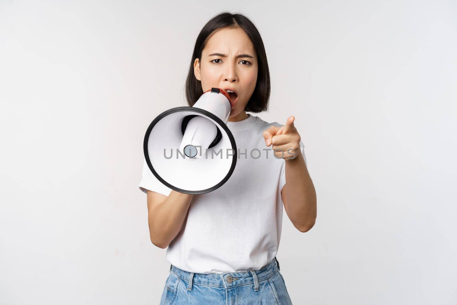 Angry asian woman with megaphone, scolding, accusing someone, protesting with speakerphone on protest, standing over white background by Benzoix