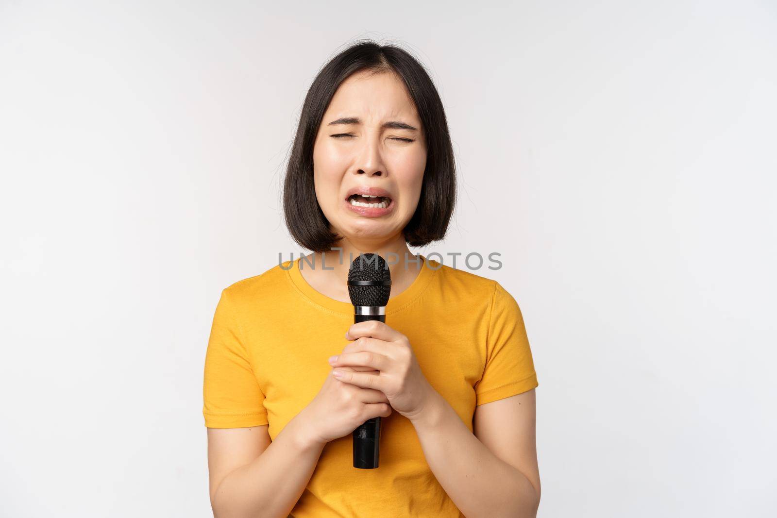 Crying asian girl singing heartbroken in microphone, holding mic and grimacing upset, standing over white background. Copy space