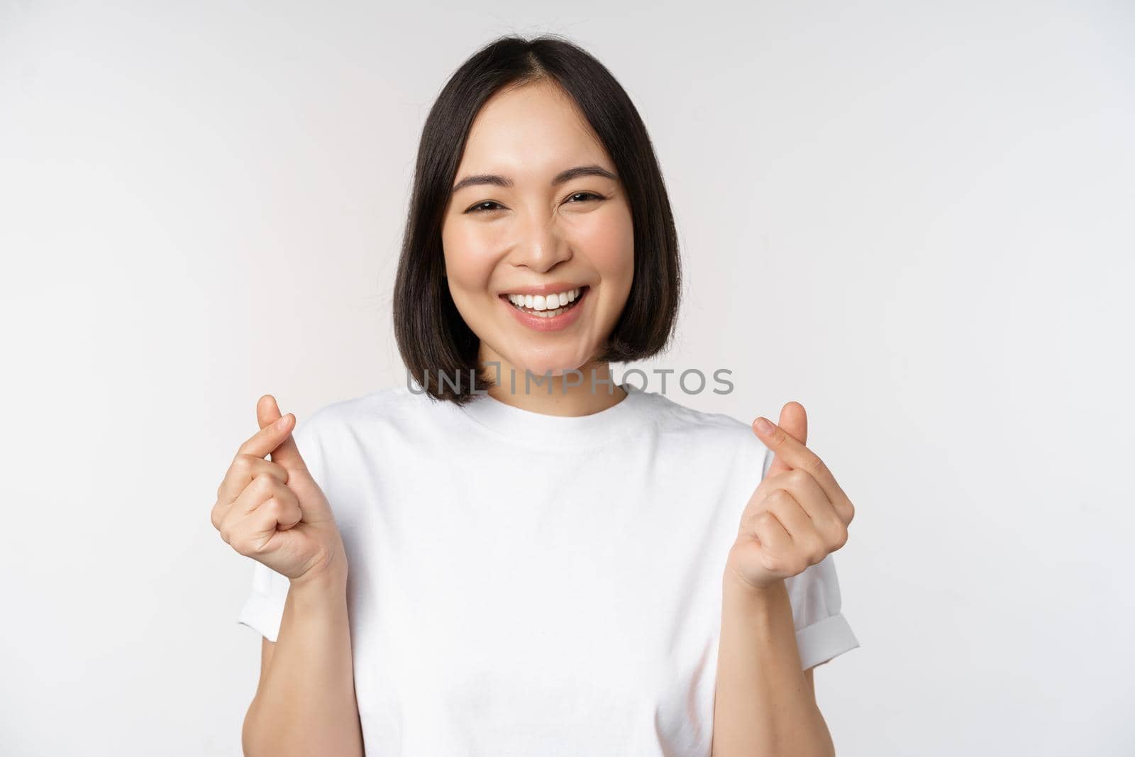 Beautiful asian woman smiling, showing finger hearts gesture, wearing tshirt, standing against white background.