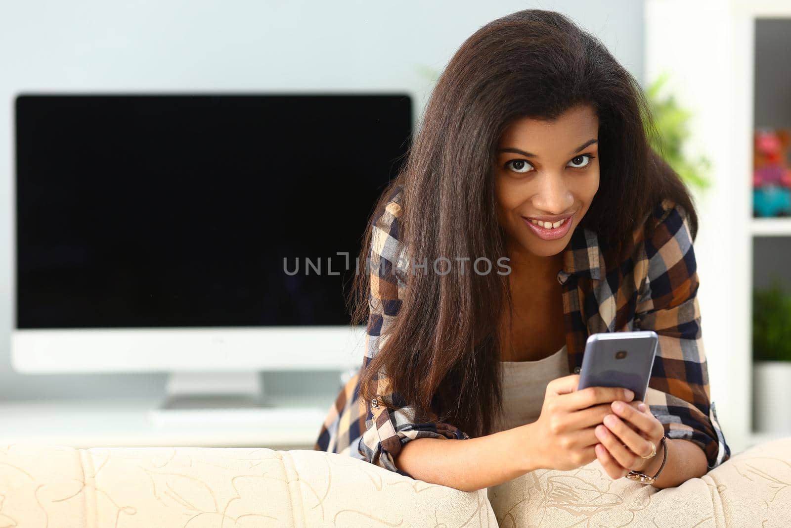 Beautiful at home by the sofa holds a smartphone, close-up. Happy african girl with device