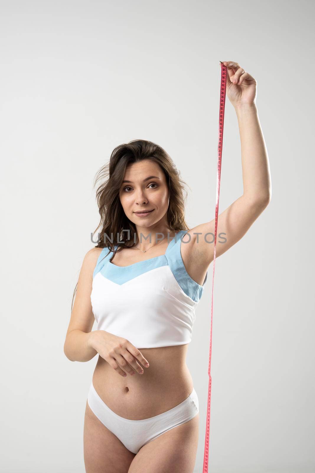 Confused brunette woman with slim body in white underwear on a diet looking in a camera and showing a pink centimeter tape. Healthy lifestyles concept. Sport and diet