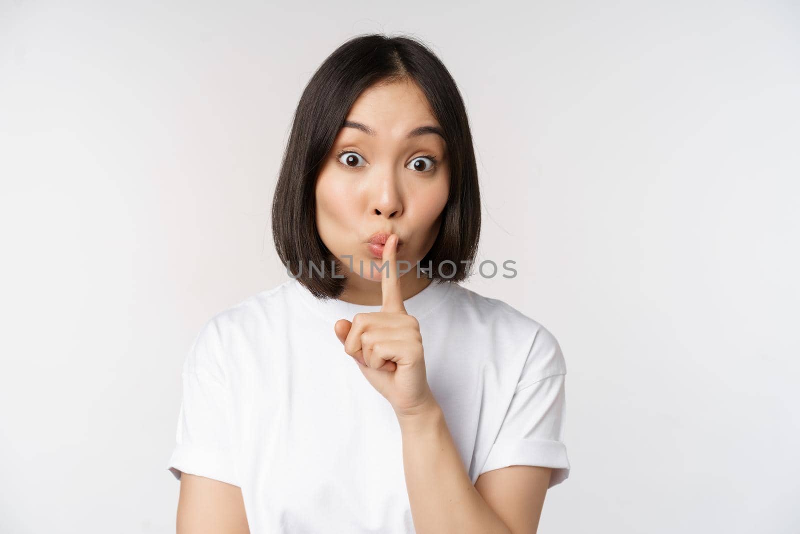 Close up portrait of young beautiful asian girl shushing, has secret, keep quiet silence gesture, press finger to lips, standing in tshirt over white background by Benzoix