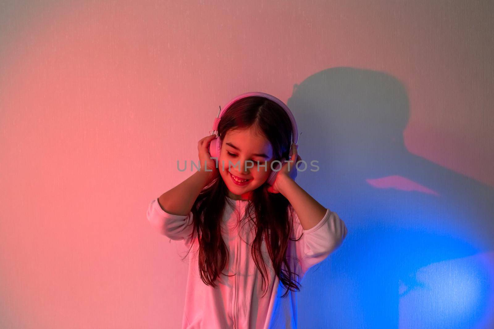 Beautiful happy little girl in pink headphones with loose dark hair listening to music in neon pink blue light holding headphones with her hands