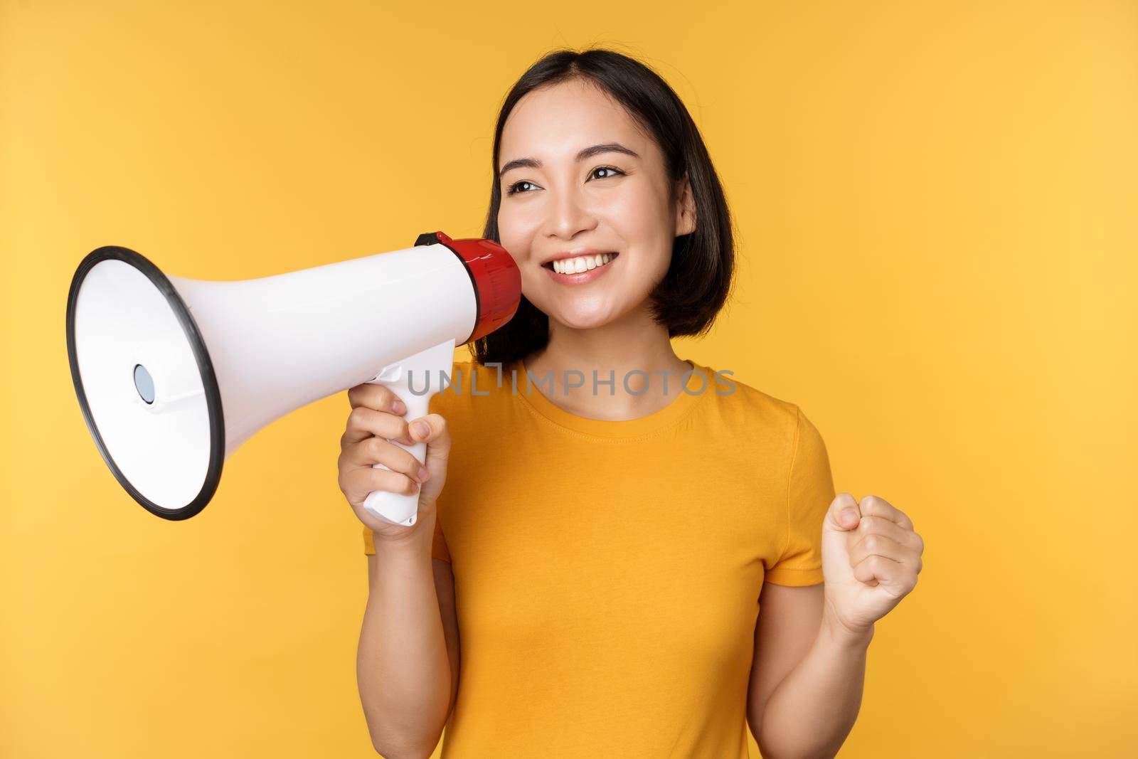 Smiling asian woman standing with megaphone, announcing smth, advertising product, standing over yellow background by Benzoix