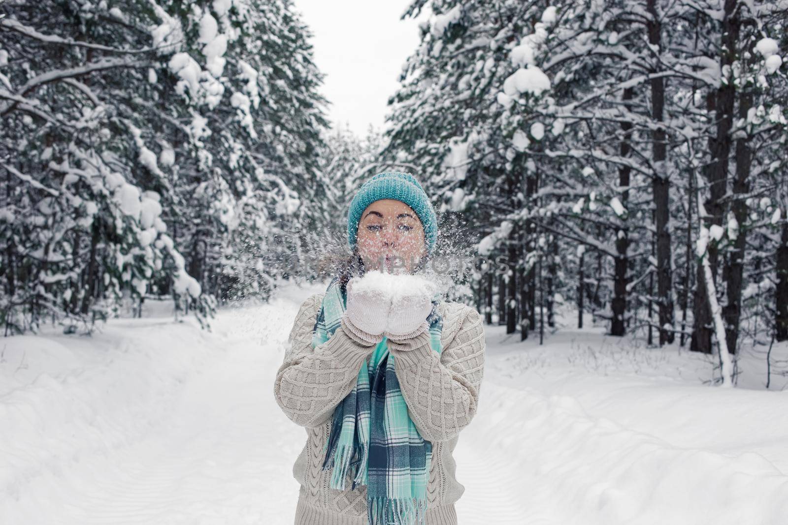 A girl in a white sweater and a light blue hat and scarf stands in a winter forest, holds snow in her hands, cheerfully blows on it