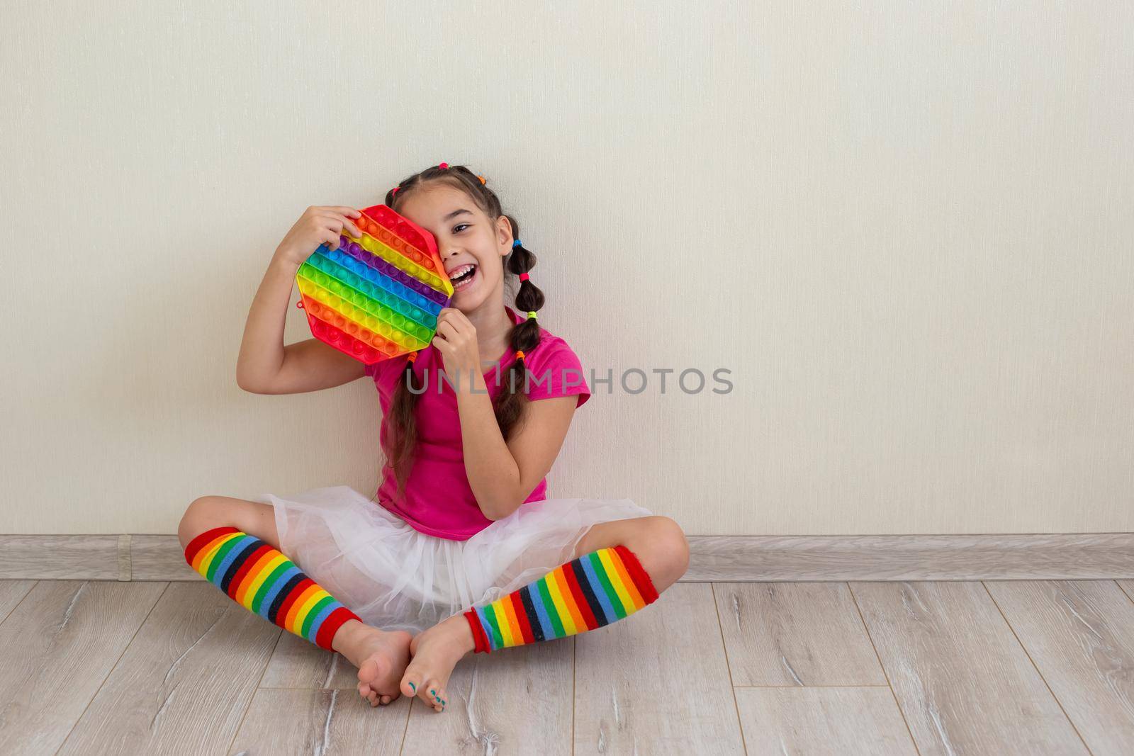 a cheerful little brunette girl in bright multicolored clothes and rainbow leggings, sits on the floor, near a light wall, holding a rainbow pop it in her hands. Copy space