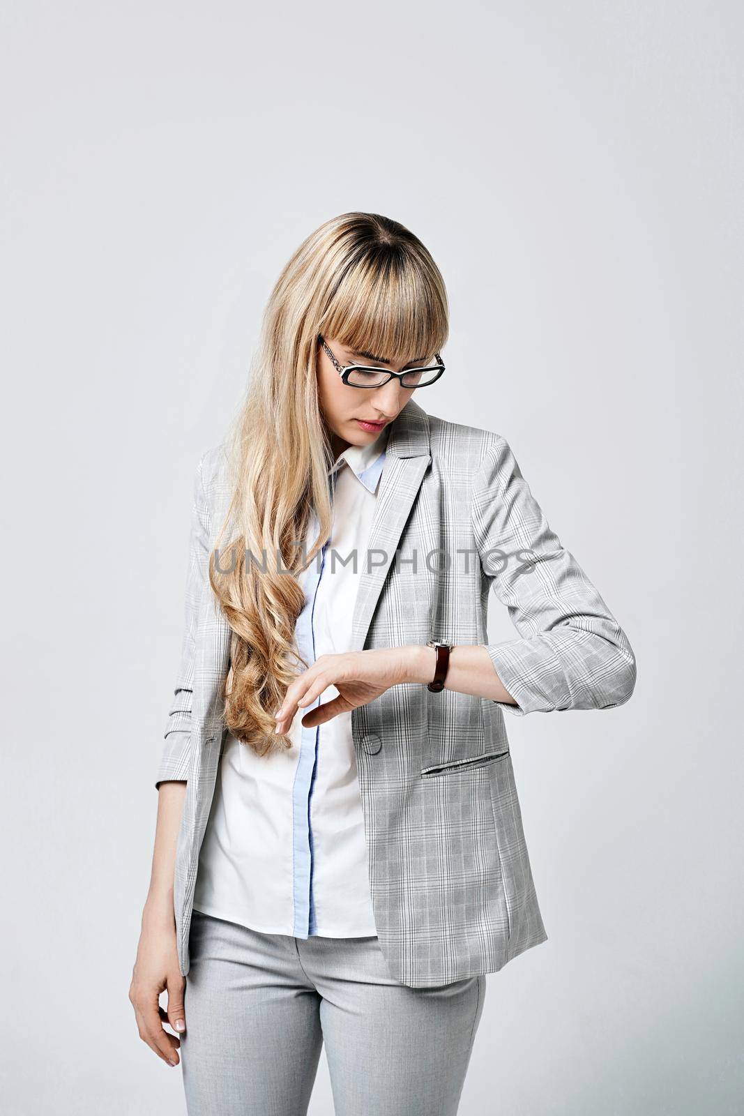 Young beautiful blonde woman in studio on light gray background. Model in pantsuit and glasses checking time with a wristwatch. by berezko
