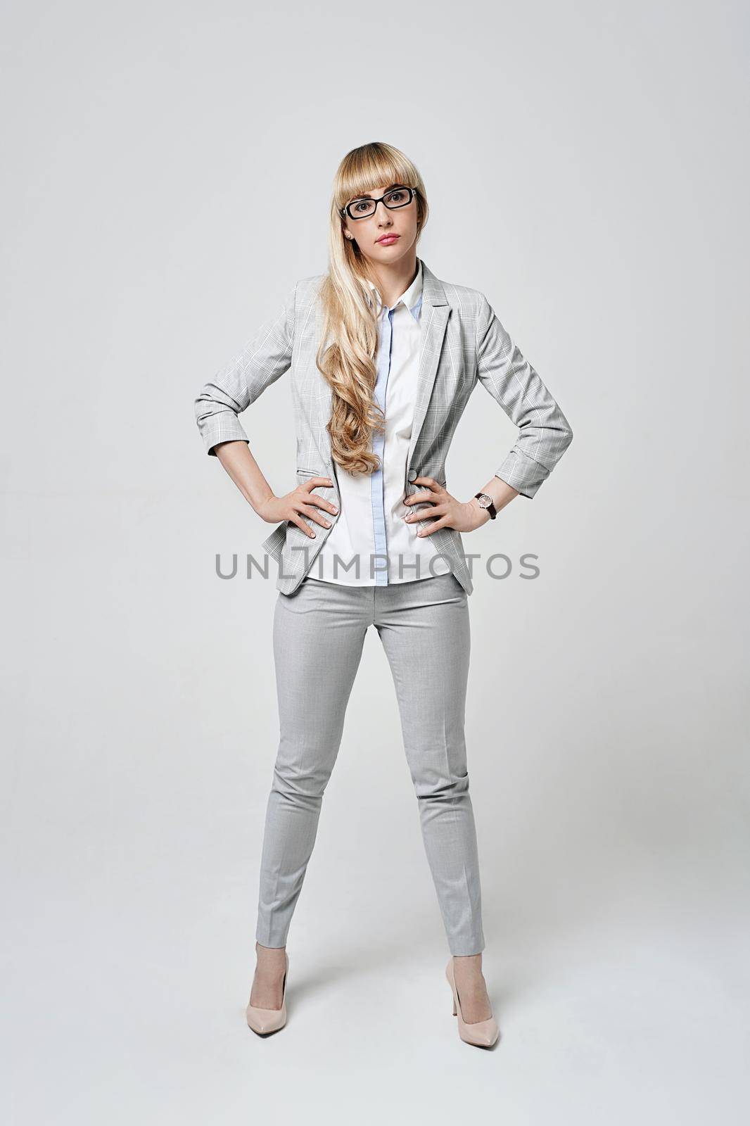 Young beautiful blonde woman in studio on light gray background. Serious model in pantsuit and glasses with arms akimbo. Dissatisfied manager. by berezko
