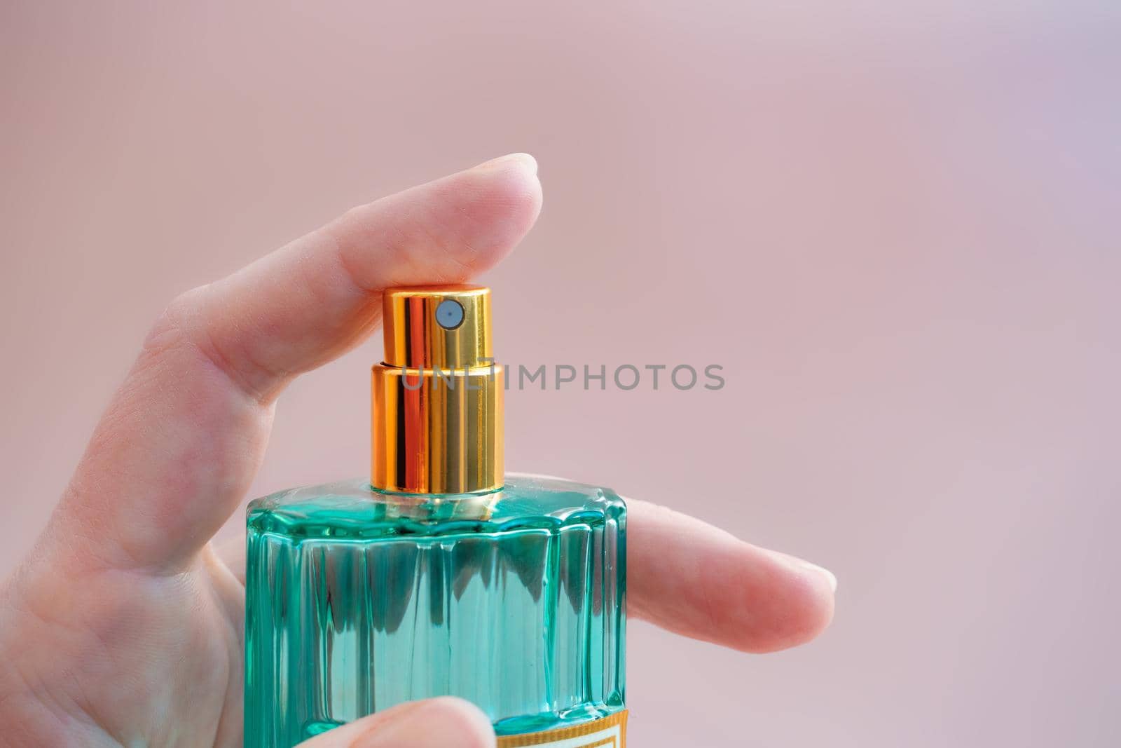 Beautiful modern perfume bottle in a female hand. Minimalistic style with transparent glass in green colors and golden lid on pink background by Andre1ns