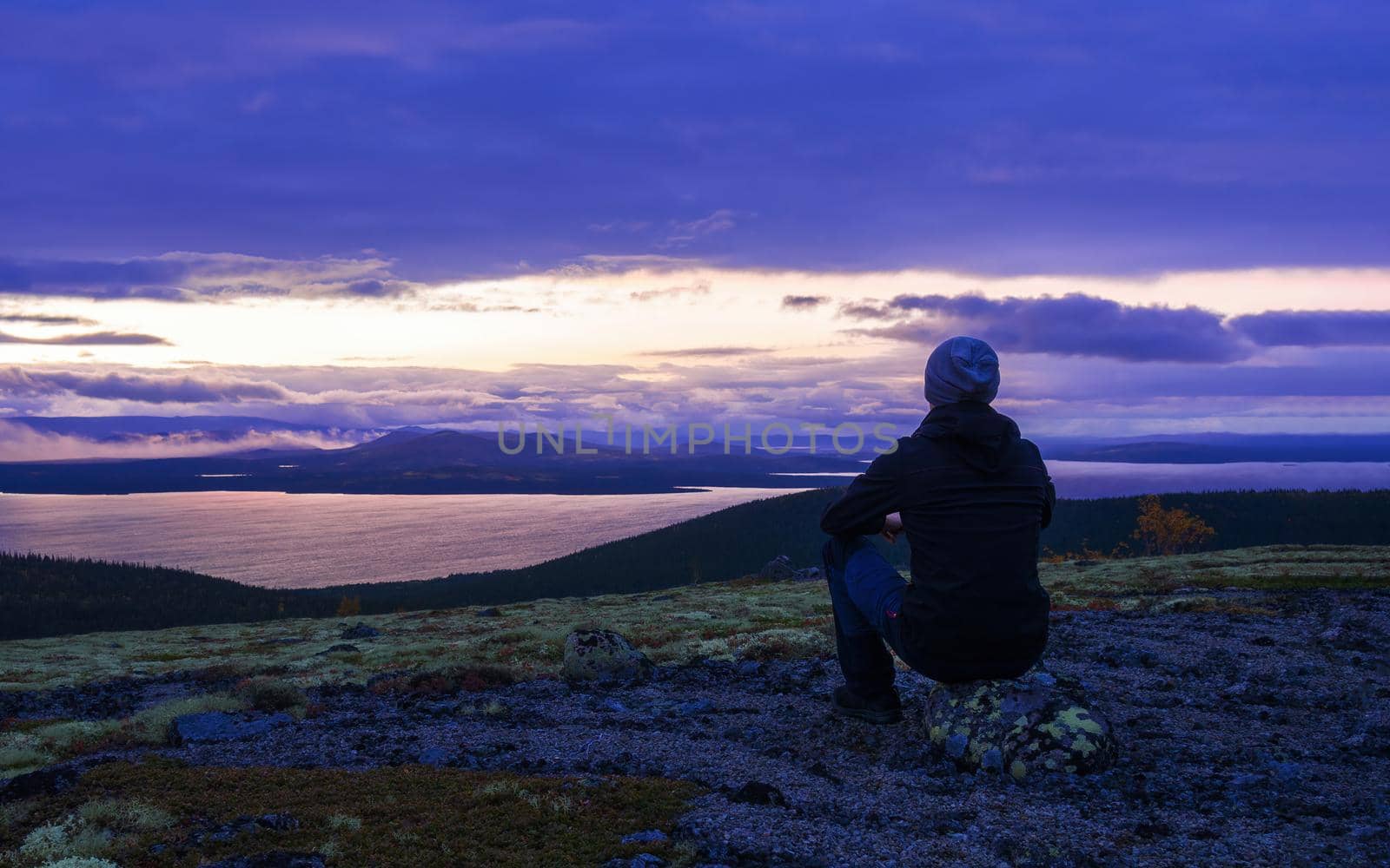 A lone tourist in a hat sits on a stone in the mountains against the backdrop of a lake at sunset, view from the back. photo