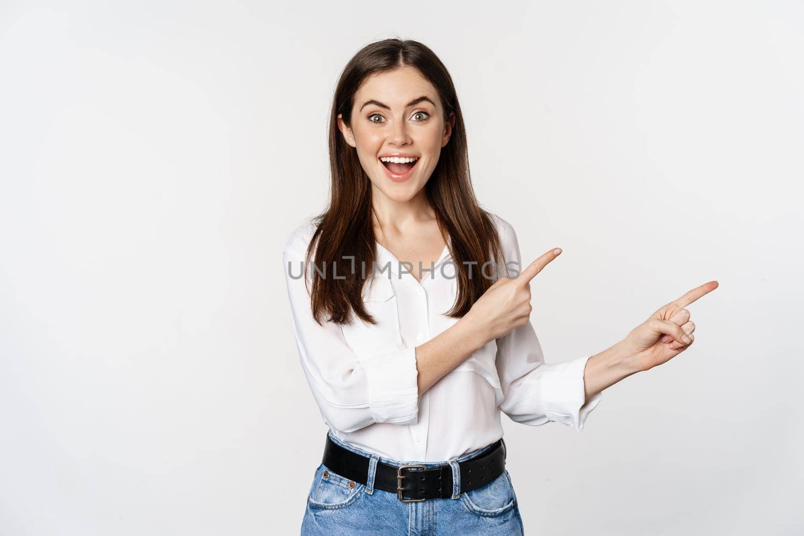Portrait of smiling confident woman, business girl showing advertisement, pointing fingers right at logo, banner or announcement, standing over white background.