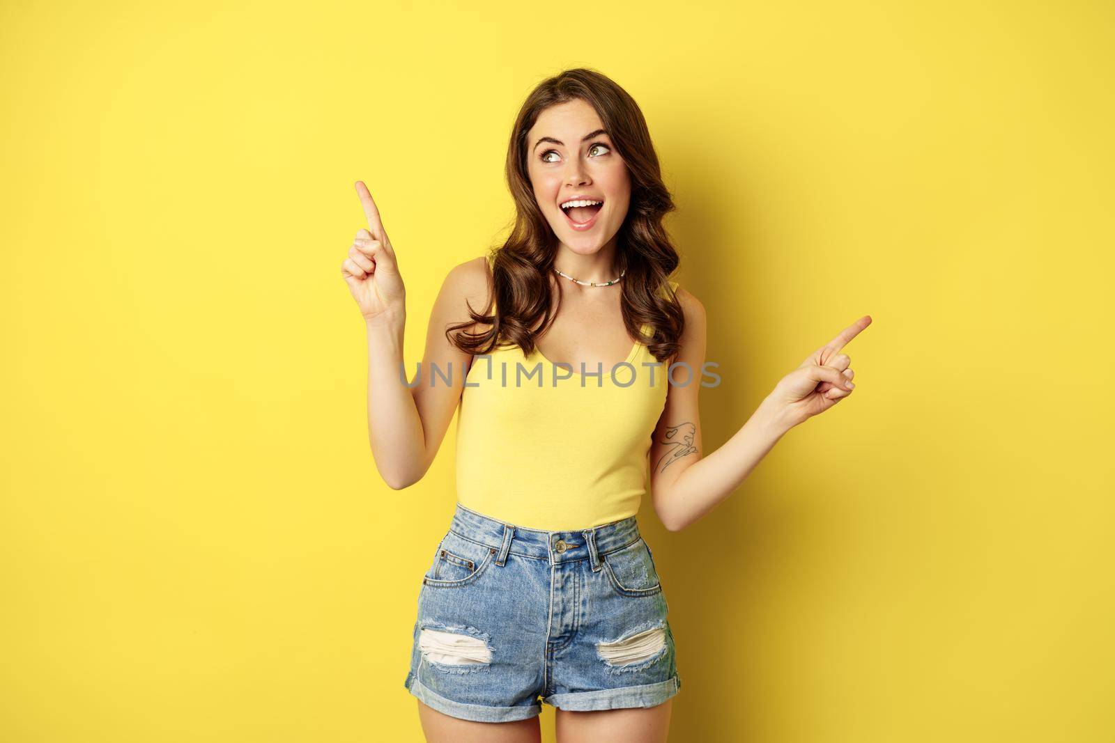 Portrait of stylish young female model, showing advertisement, pointing at logo or banner, demonstrating two ways, choices in store, standing over yellow background.