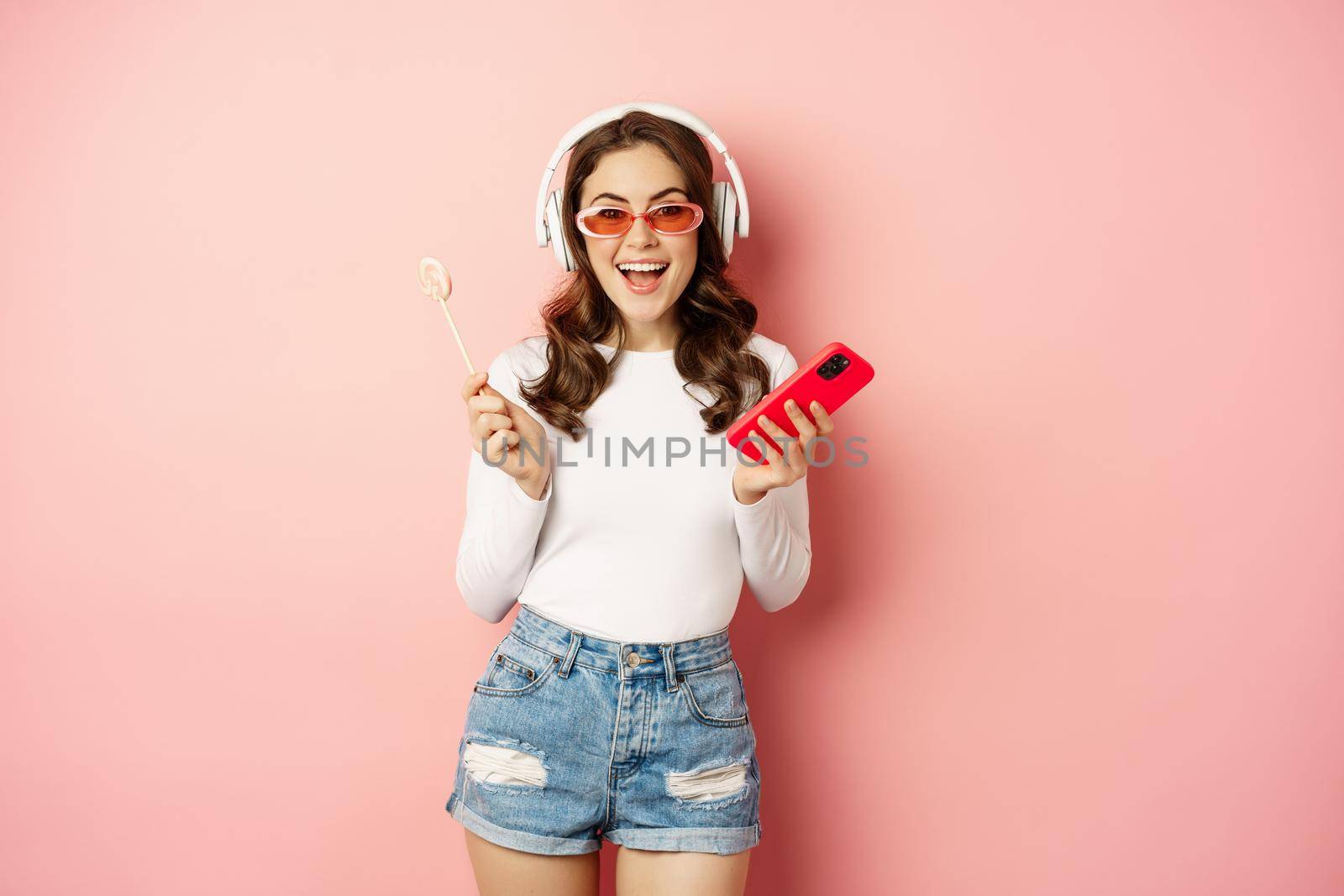 Stylish summer girl dancing with smartphone and lolipop, listening music in headphones, standing in sunglasses against pink background.