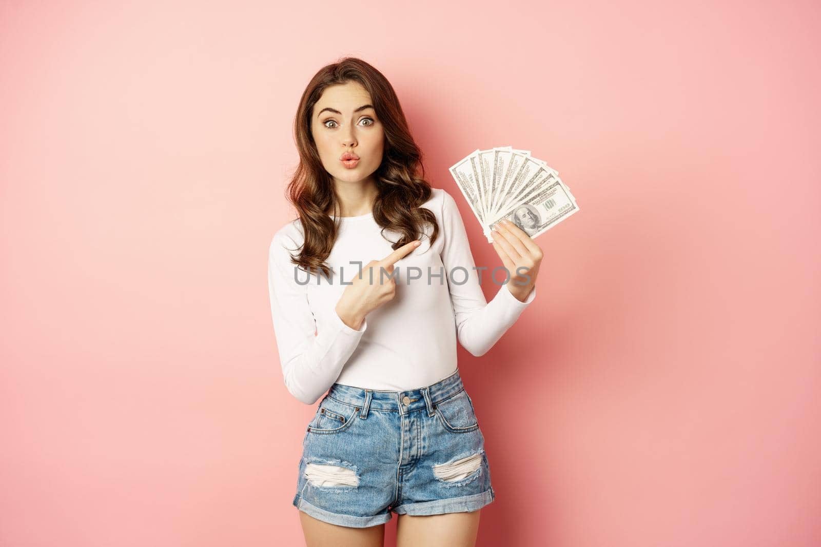 Attractive young woman holding money, cash in hands, concept of loans, microcredit and shopping, standing over pink background. Copy space