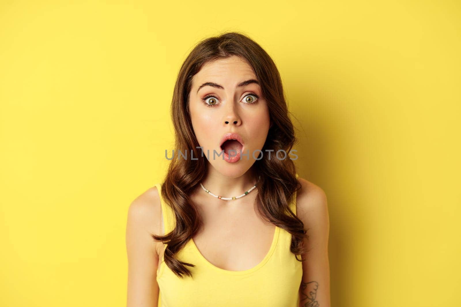 Close up portrait of stylish brunette girl looking amazed, wow face, showing excitement and surprise, impressed by something, standing over yellow background.