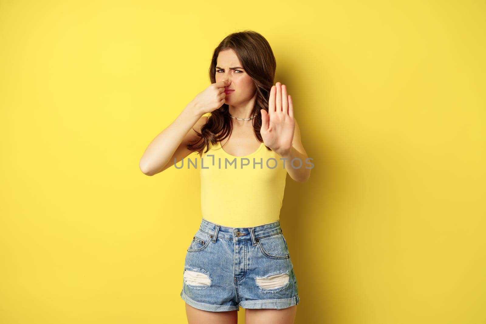 Woman showing stop sign and shut nose from disgust, dislike bad smell, declining something awful, standing over yellow background.