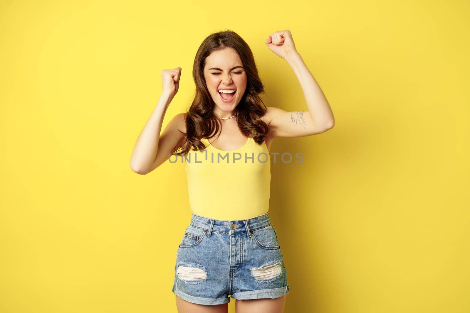 Enthusiastic young woman shouting and cheering, rejoicing, raising hands up and screaming in joy, chanting, standing over yellow background.