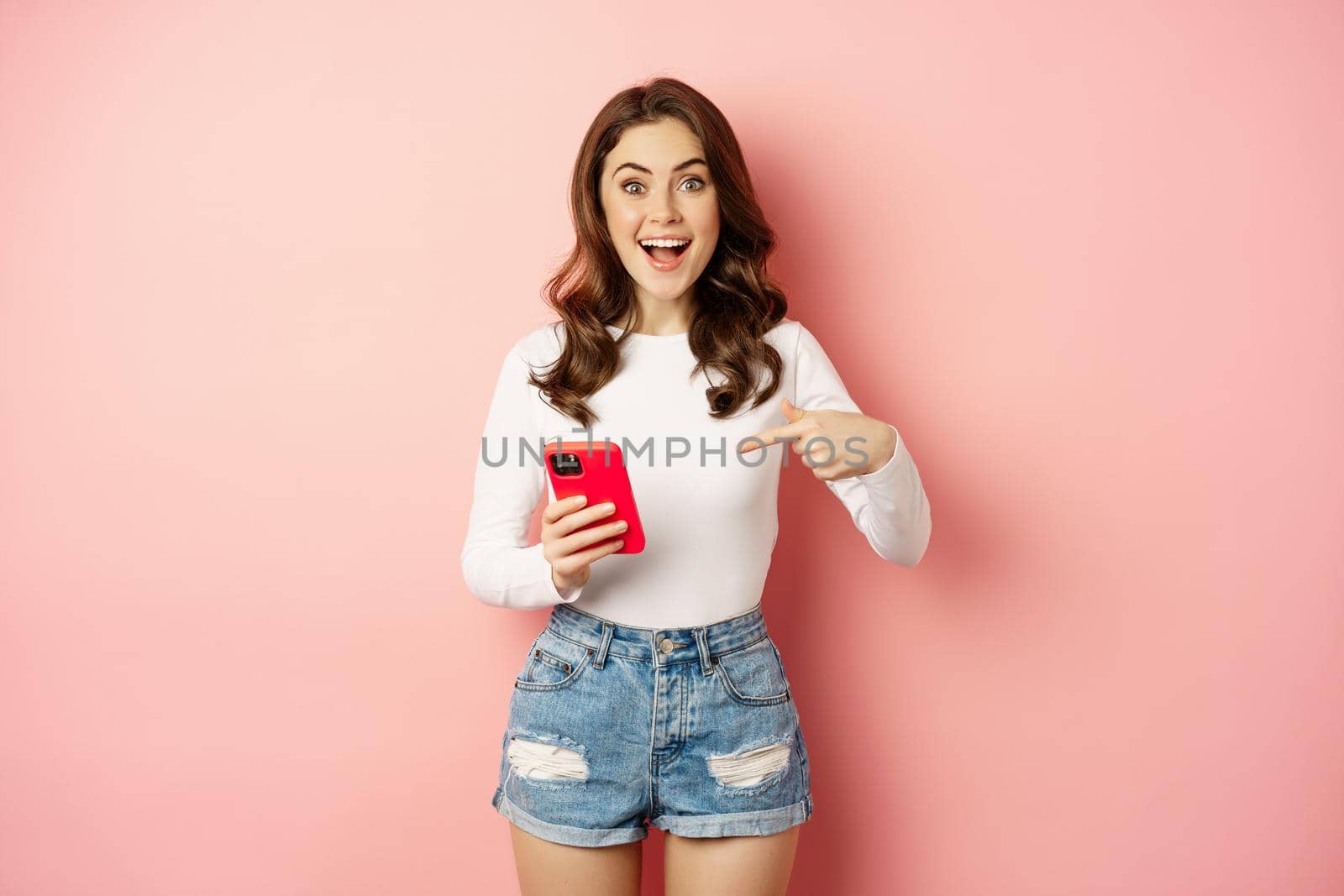 Happy brunette girl showing announcement, pointing finger at mobile phone, showing online promo, app news, standing against pink background by Benzoix