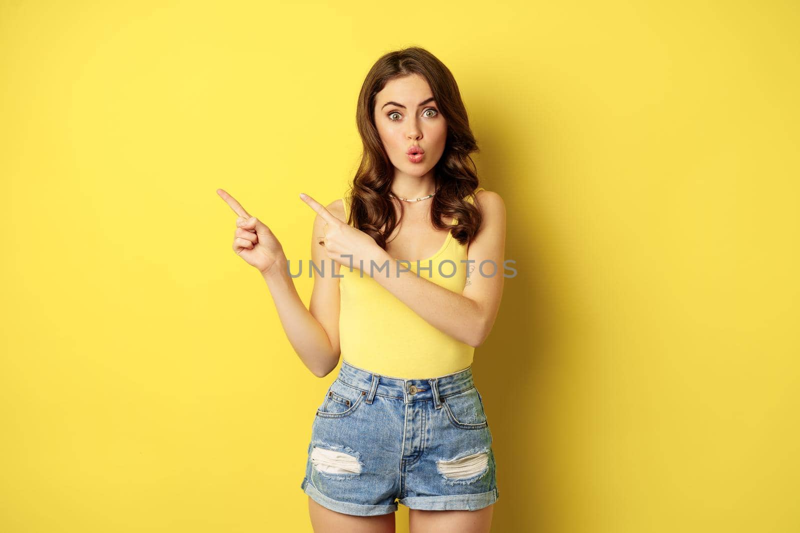 Portrait of stylish summer girl, brunette woman showing way, pointing fingers left, advertising, standing over yellow background. Copy space